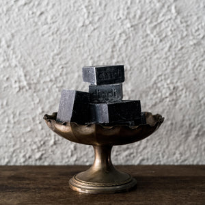 Three bars of Dindi Activated Bamboo Charcoal Soap with varying shades of grey and black, placed on a bronze-colored metal stand with a textured background.