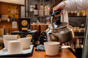A hand pouring hot water from a vintage metal kettle into a white ceramic cup on a wooden counter.